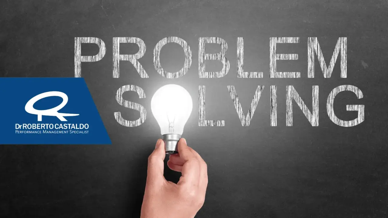 Una mano tiene una lampadina accesa, illuminando la parola "SOLVING" scritta sulla lavagna. Le parole "PROBLEM" e "SOLVING" suggeriscono l'importanza delle tecniche di problem solving.