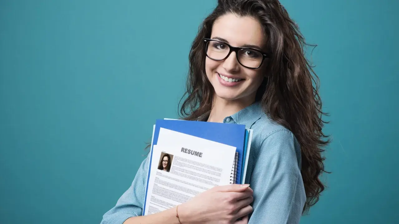 Young woman with glasses holding a resume and folders against a teal background.