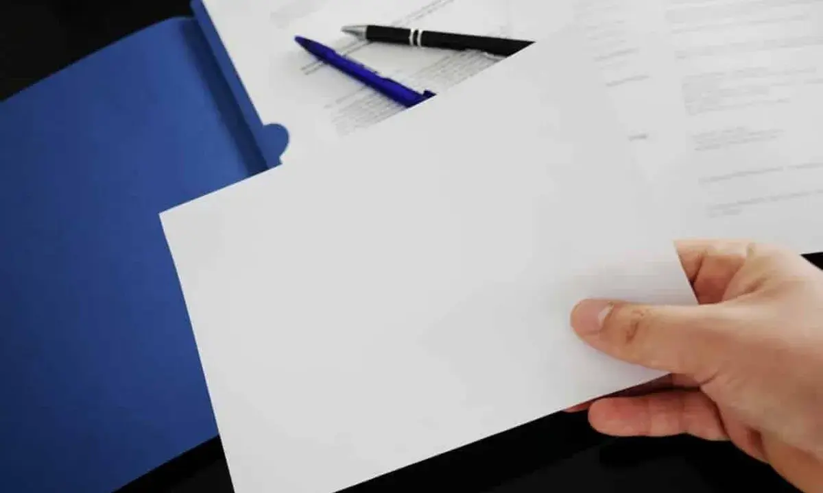 Hand holding blank paper over a blue folder with pens and documents.