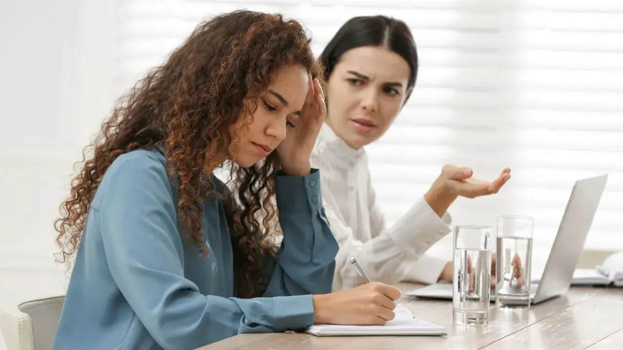 Due donne in ufficio, una con la mano sulla fronte, l'altra gesticola. Difficile gestione dei conflitti sul lavoro.