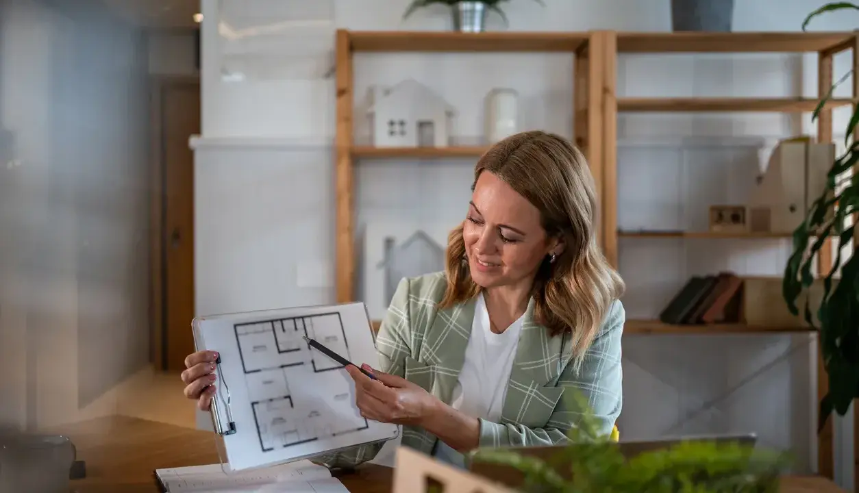 Agente immobiliare con partita IVA illustra una planimetria. L'ufficio è moderno e luminoso, con scaffali pieni di libri e oggetti decorativi.