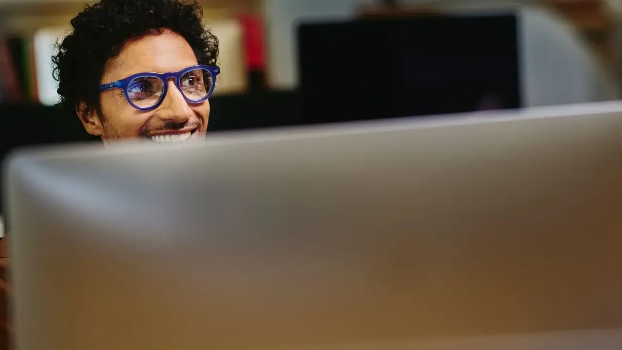 Uomo con occhiali blu sorride guardando il monitor, pensando a come migliorare le sue competenze digitali per il CV.