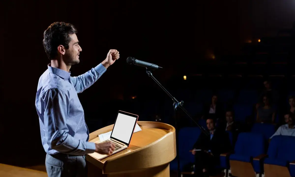Un oratore sul palco, con microfono e laptop, illustra cos'è il public speaking con un gesto enfatico.