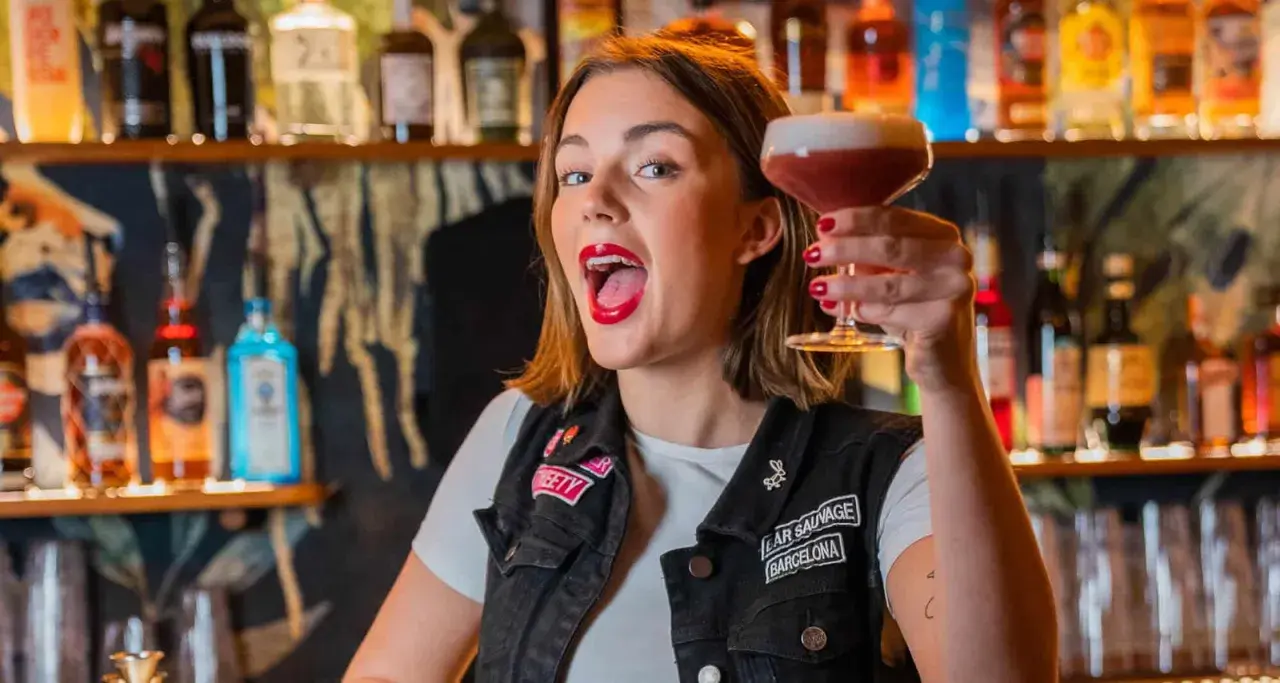 Giovane donna con rossetto rosso acceso, in un bar, solleva un cocktail. Le sue attitudini sono un esempio di gioia e vivacità.