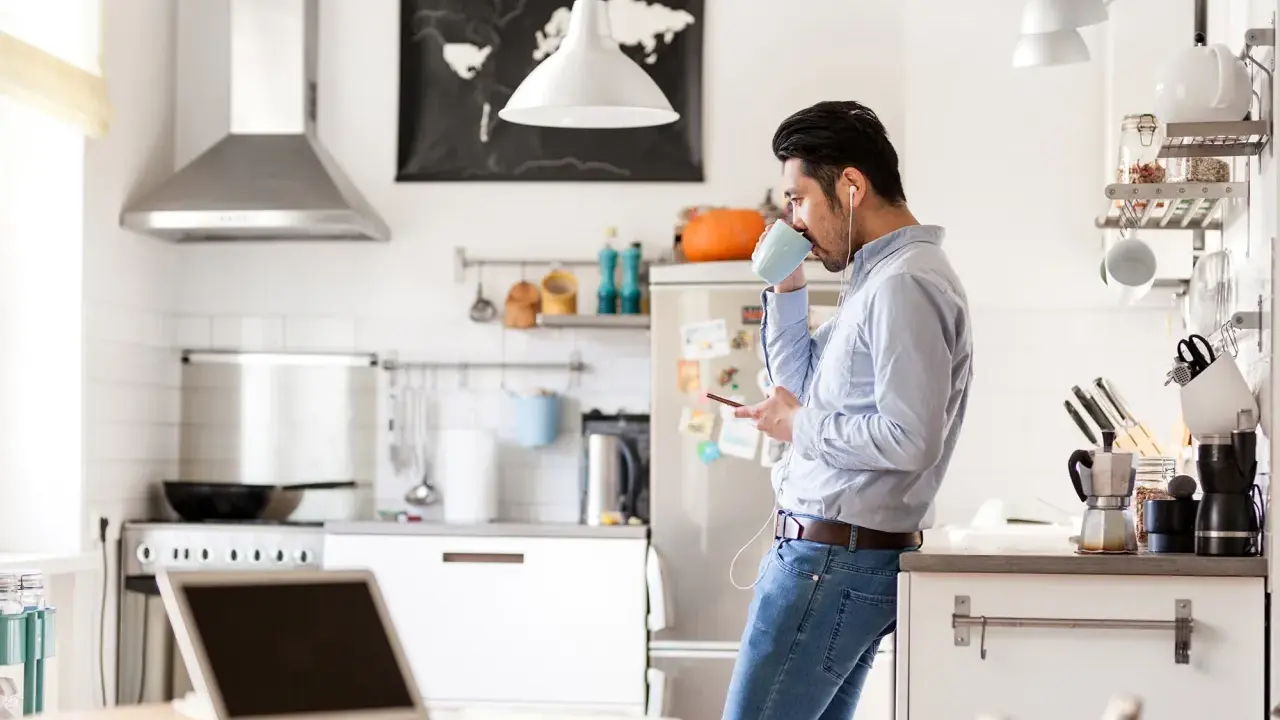 Uomo in cucina con caffè e smartphone, laptop aperto sul tavolo. Un esempio di **modalità ibrida** tra lavoro e vita domestica.