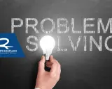 Una mano tiene una lampadina accesa, illuminando la parola "SOLVING" scritta sulla lavagna. Le parole "PROBLEM" e "SOLVING" suggeriscono l'importanza delle tecniche di problem solving.