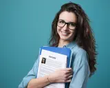 Young woman with glasses holding a resume and folders against a teal background.