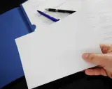 Hand holding blank paper over a blue folder with pens and documents.