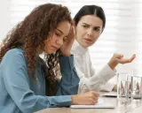 Due donne in ufficio, una con la mano sulla fronte, l'altra gesticola. Difficile gestione dei conflitti sul lavoro.
