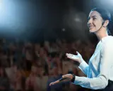 Donna sorridente con microfono, sul palco davanti a un pubblico. Esercizi per parlare in pubblico efficaci.
