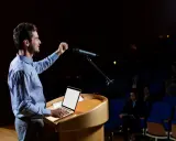 Un oratore sul palco, con microfono e laptop, illustra cos'è il public speaking con un gesto enfatico.