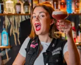 Giovane donna con rossetto rosso acceso, in un bar, solleva un cocktail. Le sue attitudini sono un esempio di gioia e vivacità.