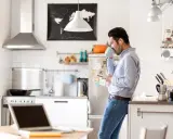 Uomo in cucina con caffè e smartphone, laptop aperto sul tavolo. Un esempio di **modalità ibrida** tra lavoro e vita domestica.