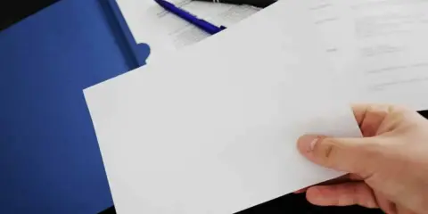 Hand holding blank paper over a blue folder with pens and documents.