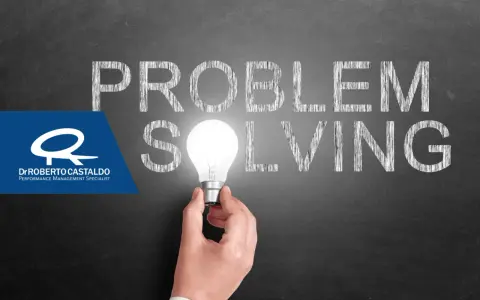 Una mano tiene una lampadina accesa, illuminando la parola "SOLVING" scritta sulla lavagna. Le parole "PROBLEM" e "SOLVING" suggeriscono l'importanza delle tecniche di problem solving.