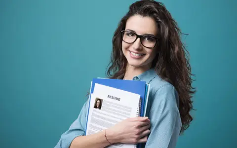Young woman with glasses holding a resume and folders against a teal background.