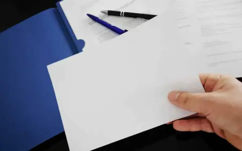 Hand holding blank paper over a blue folder with pens and documents.