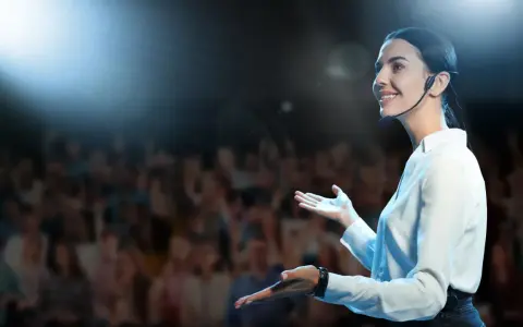 Donna sorridente con microfono, sul palco davanti a un pubblico. Esercizi per parlare in pubblico efficaci.