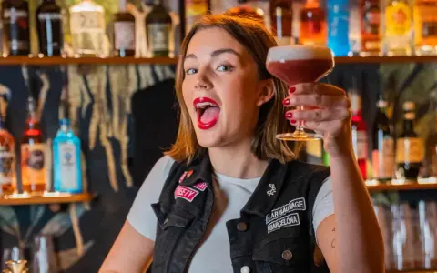 Giovane donna con rossetto rosso acceso, in un bar, solleva un cocktail. Le sue attitudini sono un esempio di gioia e vivacità.