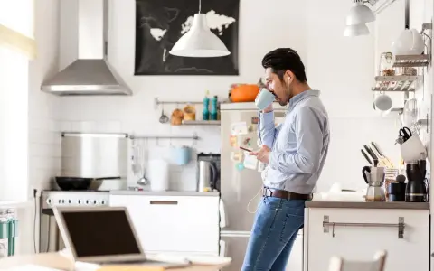 Uomo in cucina con caffè e smartphone, laptop aperto sul tavolo. Un esempio di **modalità ibrida** tra lavoro e vita domestica.