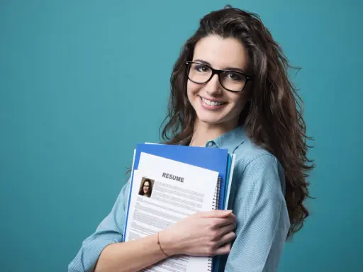 Young woman with glasses holding a resume and folders against a teal background.