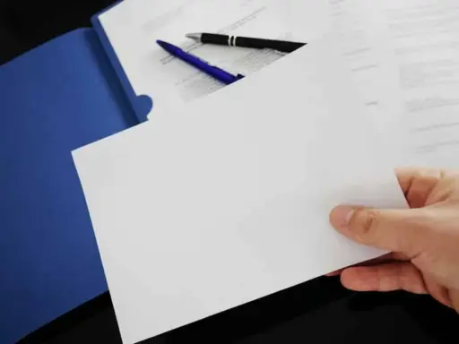 Hand holding blank paper over a blue folder with pens and documents.
