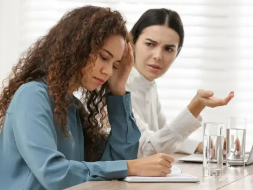 Due donne in ufficio, una con la mano sulla fronte, l'altra gesticola. Difficile gestione dei conflitti sul lavoro.