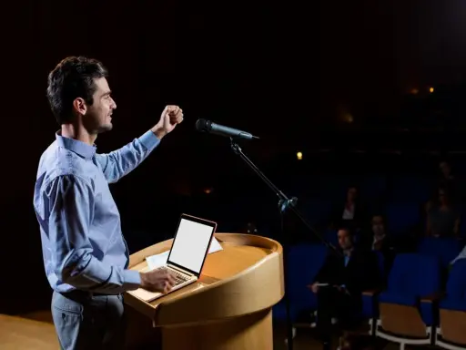 Un oratore sul palco, con microfono e laptop, illustra cos'è il public speaking con un gesto enfatico.