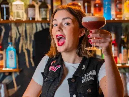 Giovane donna con rossetto rosso acceso, in un bar, solleva un cocktail. Le sue attitudini sono un esempio di gioia e vivacità.