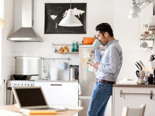 Uomo in cucina con caffè e smartphone, laptop aperto sul tavolo. Un esempio di **modalità ibrida** tra lavoro e vita domestica.