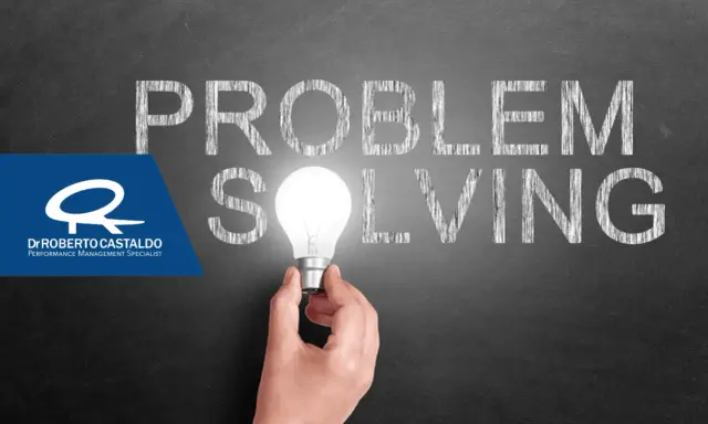 Una mano tiene una lampadina accesa, illuminando la parola "SOLVING" scritta sulla lavagna. Le parole "PROBLEM" e "SOLVING" suggeriscono l'importanza delle tecniche di problem solving.