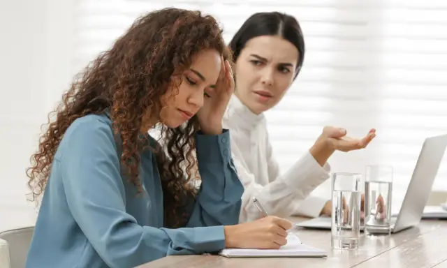Due donne in ufficio, una con la mano sulla fronte, l'altra gesticola. Difficile gestione dei conflitti sul lavoro.