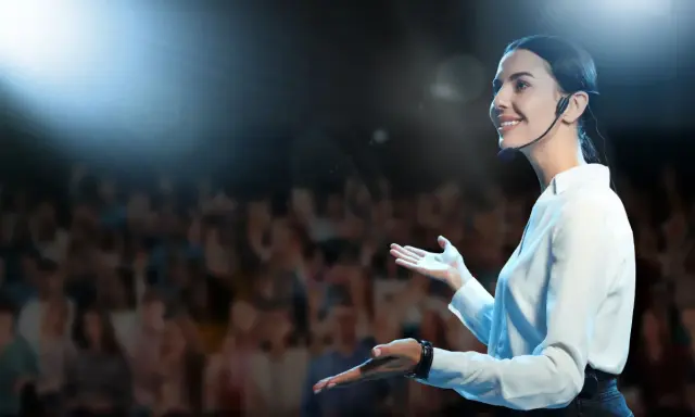 Donna sorridente con microfono, sul palco davanti a un pubblico. Esercizi per parlare in pubblico efficaci.