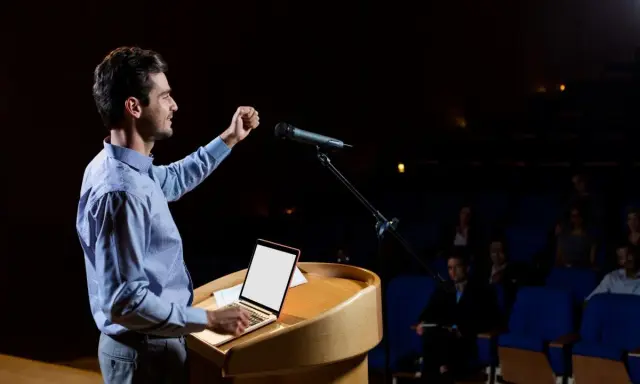Un oratore sul palco, con microfono e laptop, illustra cos'è il public speaking con un gesto enfatico.