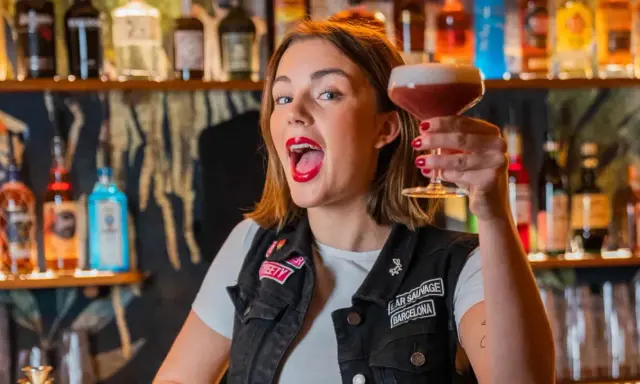 Giovane donna con rossetto rosso acceso, in un bar, solleva un cocktail. Le sue attitudini sono un esempio di gioia e vivacità.