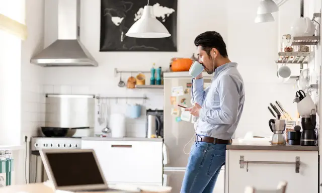 Uomo in cucina con caffè e smartphone, laptop aperto sul tavolo. Un esempio di **modalità ibrida** tra lavoro e vita domestica.