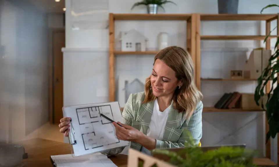 Agente immobiliare con partita IVA illustra una planimetria. L'ufficio è moderno e luminoso, con scaffali pieni di libri e oggetti decorativi.