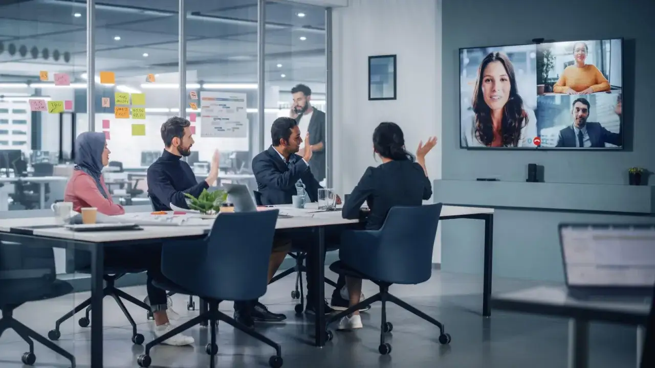 Team in riunione, alcuni in presenza, altri in videochiamata su un grande schermo, per una perfetta modalit&agrave; ibrida di lavoro.