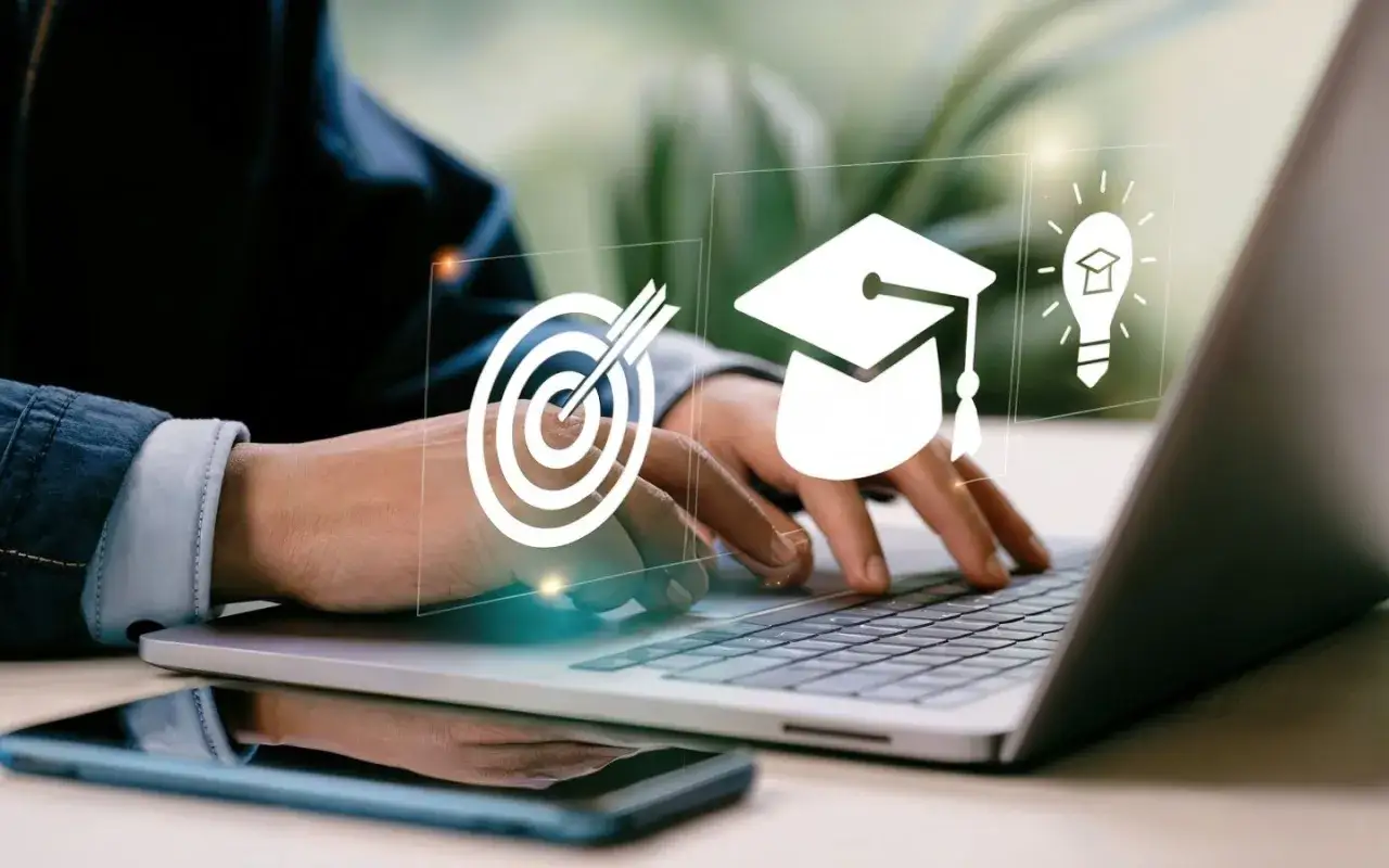 Mani che digitano su un laptop, con icone di un bersaglio, un cappello da laureato e una lampadina che simboleggiano il raggiungimento di obiettivi e l'apprendimento. Scopri il google digital training gratis per la tua crescita.